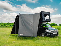 Small black car with the TentBox GO and Tunnel Awning set up in a grassy field under a blue sky with clouds.